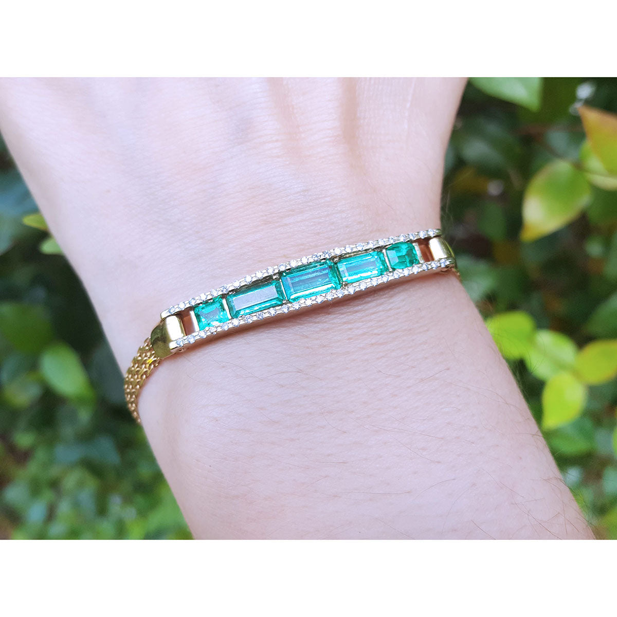 Gold bracelet with vife genuine emeralds on a wrist against a blurred green background.