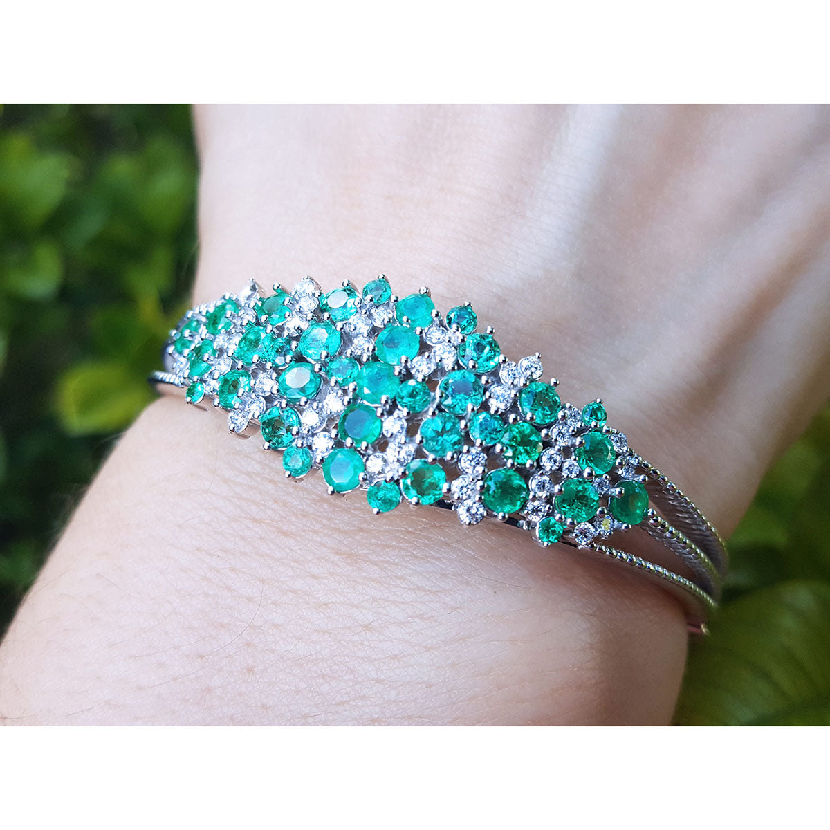 A close-up view of a bangle bracelet with emerald and diamonds on a wrist against a blurred green background.