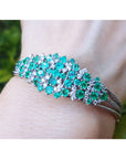 A close-up view of a bangle bracelet with emerald and diamonds on a wrist against a blurred green background.