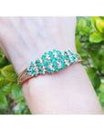 On a wrist a gold bangle bracelet with colombian emeralds and diamonds on a green leafy background.