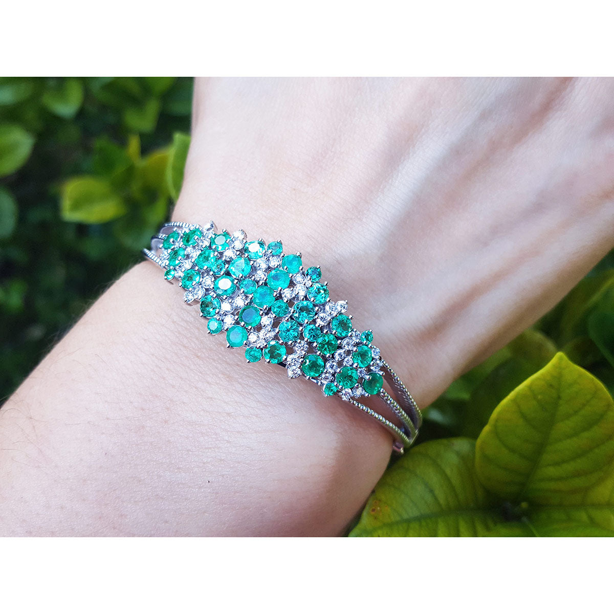 A white gold emerald bangle bracelet on a wrist against a green leafy background.