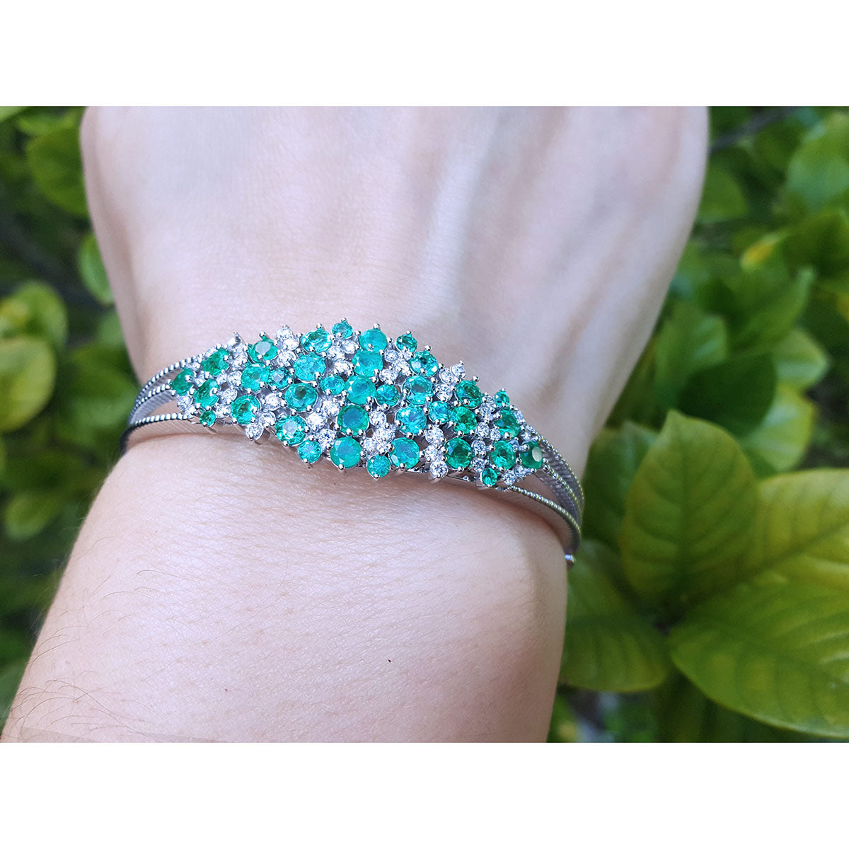 Bracelet with round cut emerald and diamonds on a wrist against a green leafy background.