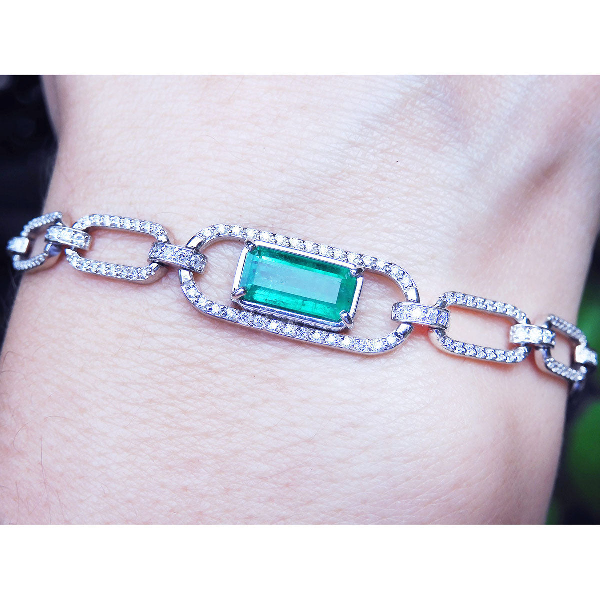 White and yellow gold paperclip bracelet with an emerald-cut natural emerald on a wrist against a neutral background.