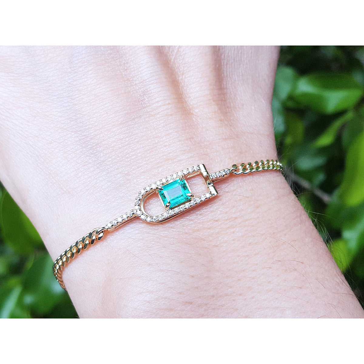 Gold bracelet with a colombian emerald on a wrist against a blurred green background.