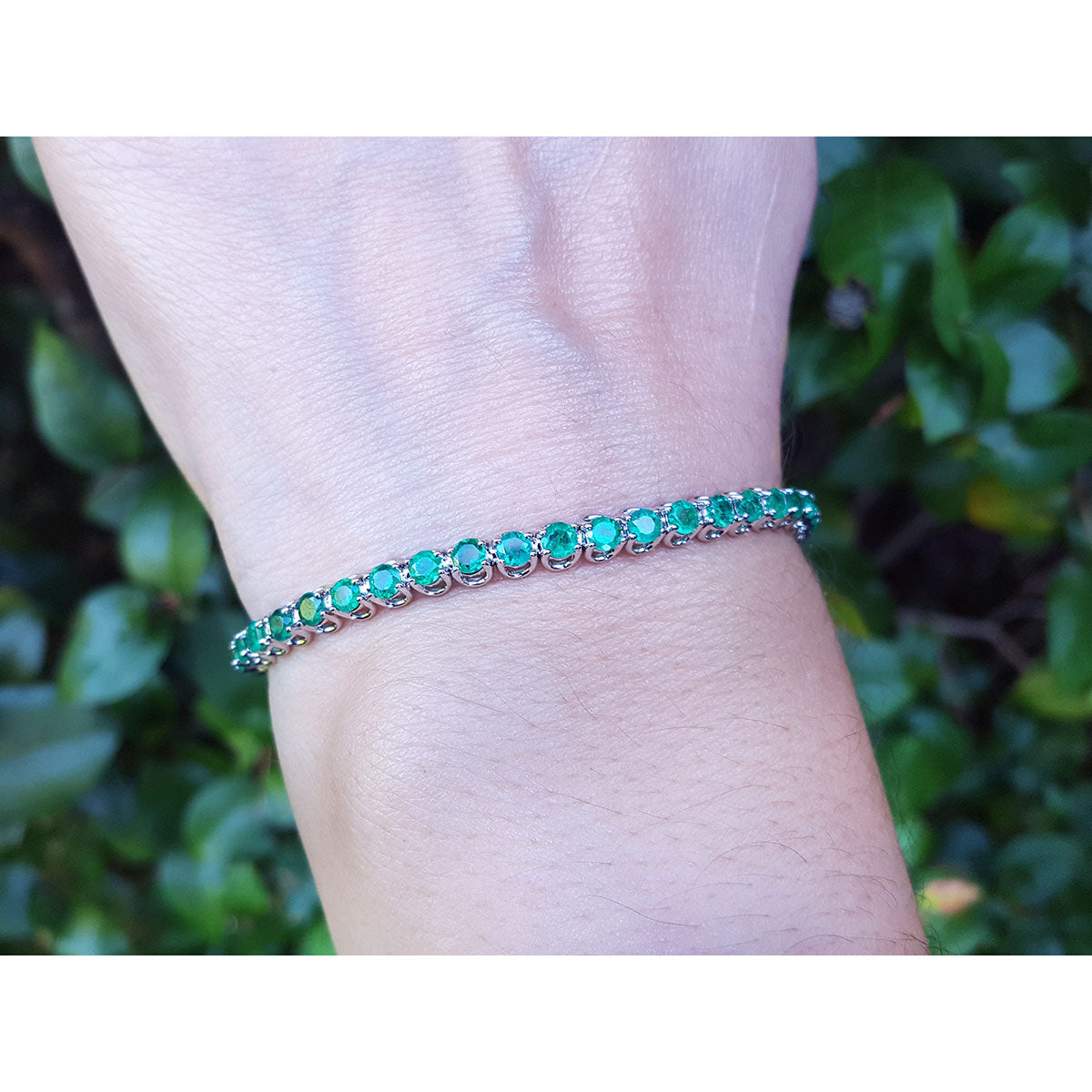 A natural colombian emerald tennis bracelet on a wrist against a blurred green background.
