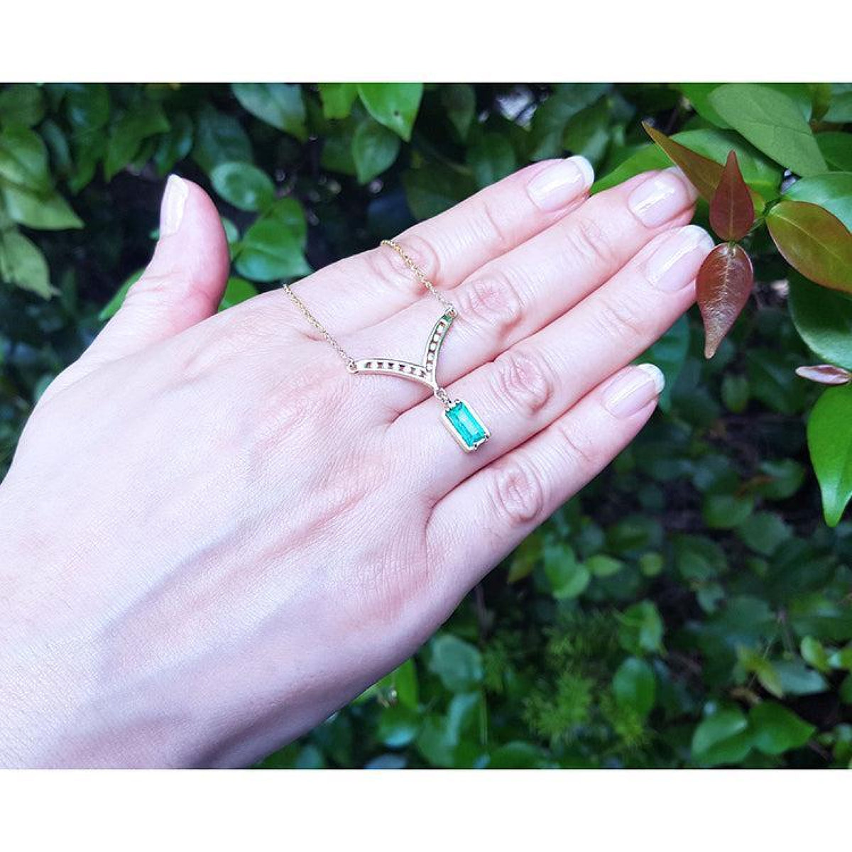 14K yellow gold necklace with a baguette-cut Colombian emerald dangling from a diamond-accented V-pendant, shown worn on a person's finger with their hand resting against a background of green leaves.