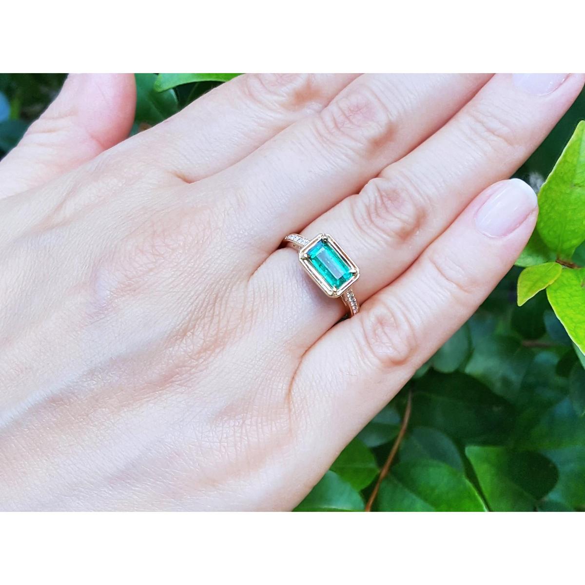 Hand wearing a ring with an emerald stone against a green leafy background.