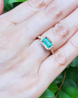 Hand wearing a ring with an emerald stone against a green leafy background.