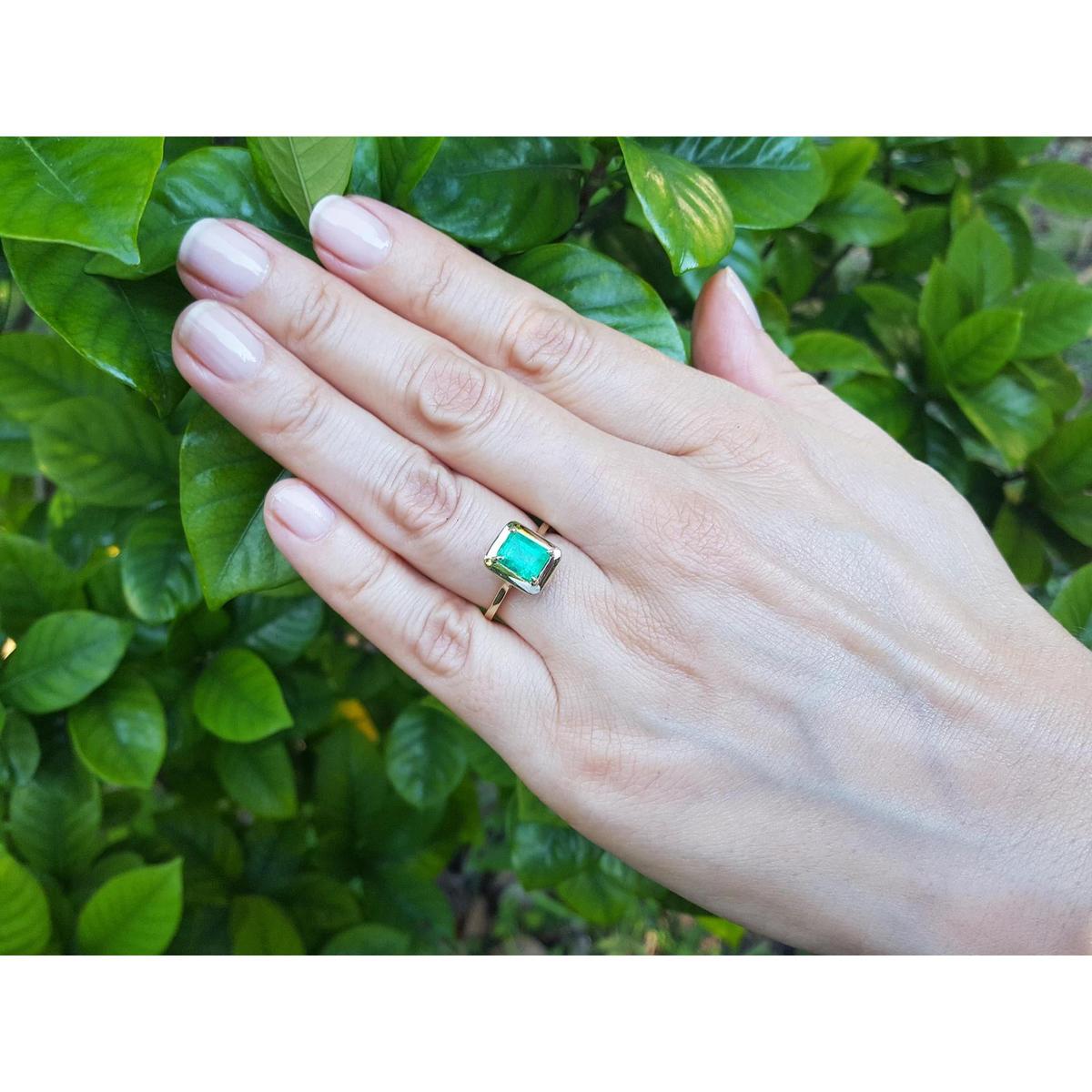 A hand wearing an emerald solitaire ring yellow gold against a green leafy background.