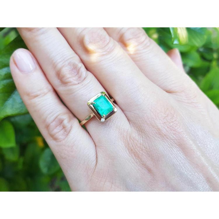 A close-up of a hand wearing a yellow gold emerald solitaire ring on a green blurry background.
