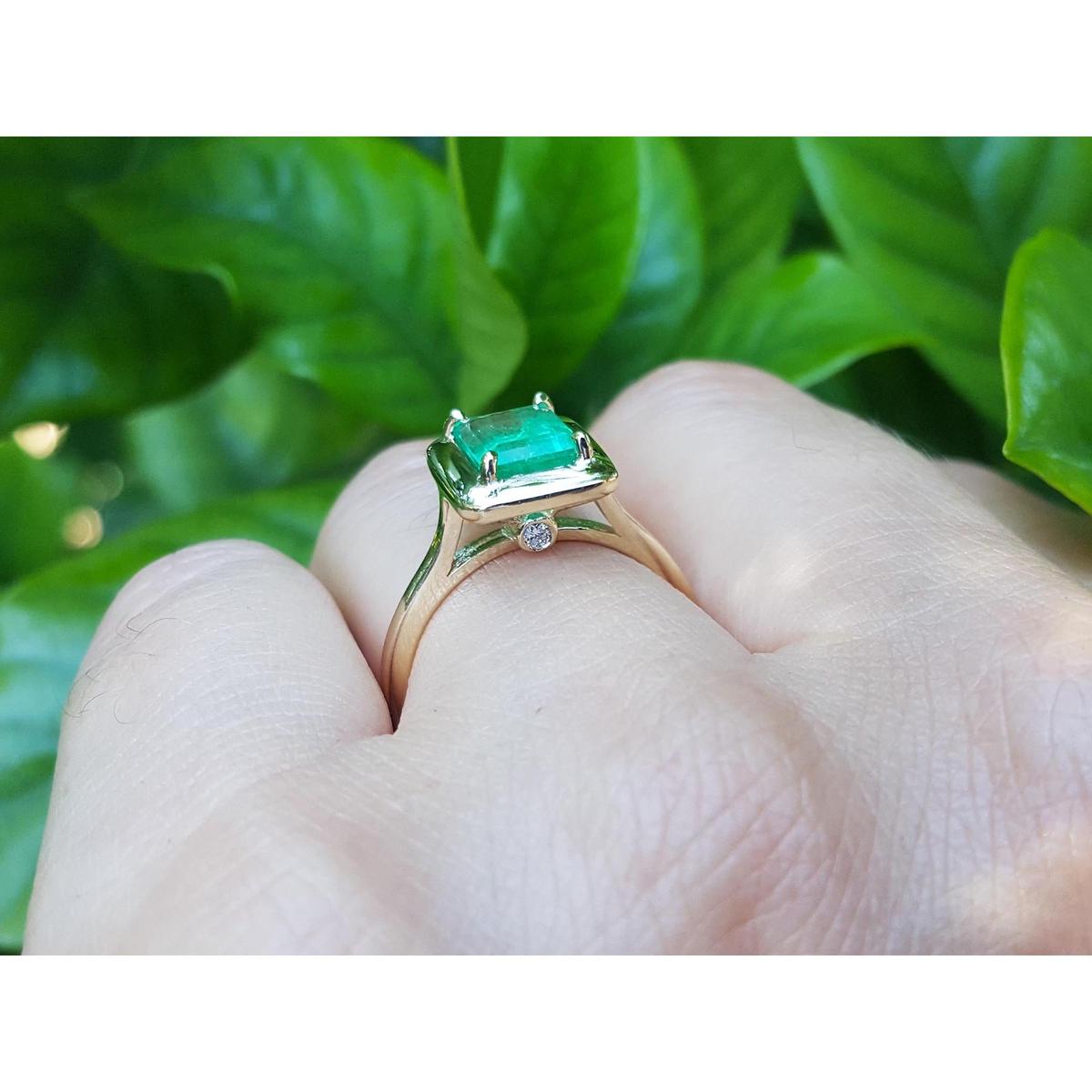 On a hand the underside view of an emerald solitaire ring in yellow gold against a leafy background.