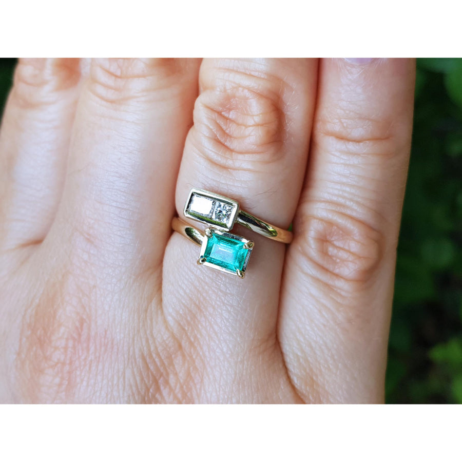 A gold ring with a rectangular clear gemstone and an emerald-cut turquoise green gemstone, shown worn on a person's finger.