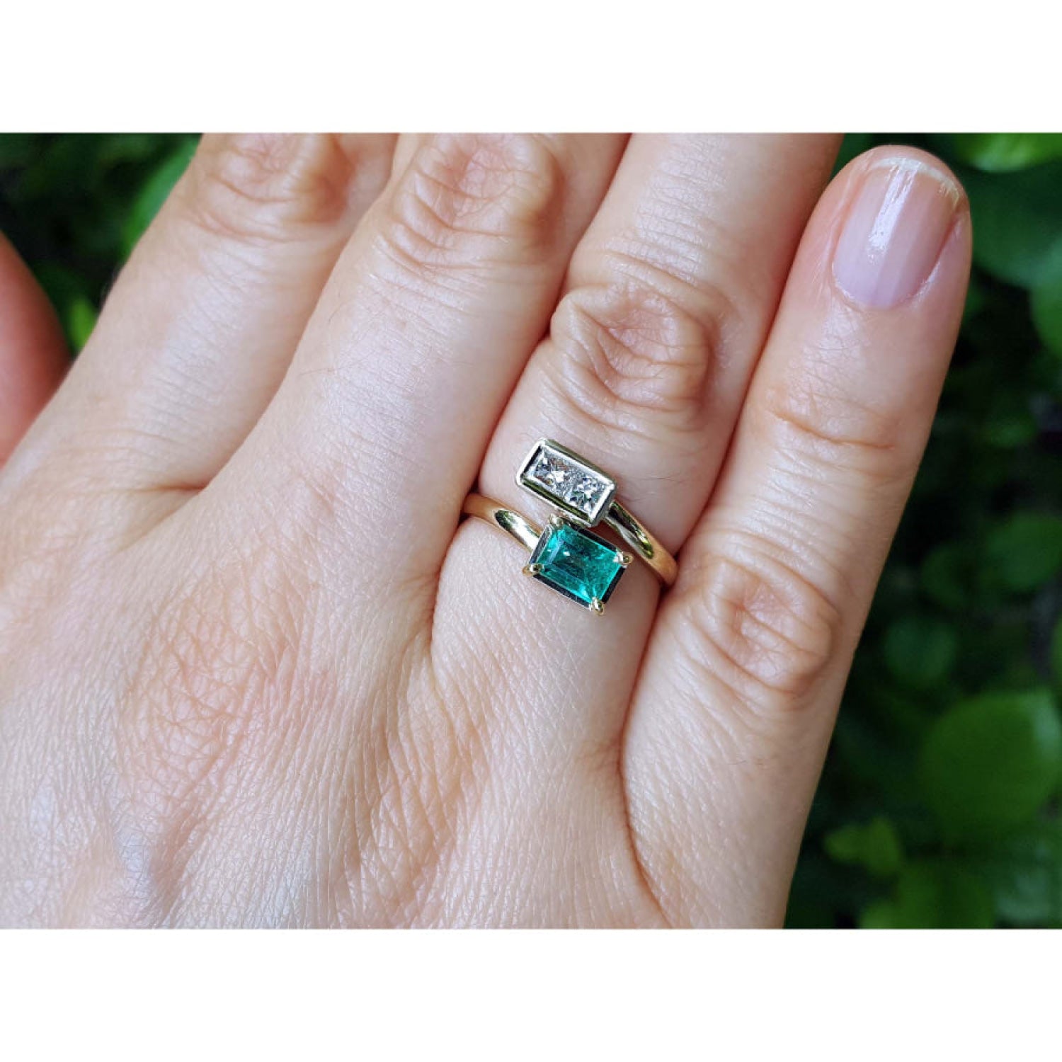 A close-up of a hand wearing a 18k yellow gold toi et moi emerald and diamond ring, against a blurred green background.