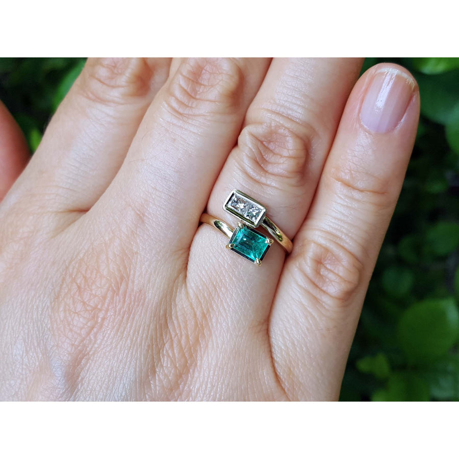 A close-up of a hand wearing a gold ring set with a rectangular clear gemstone and a larger rectangular emerald-cut green gemstone, against a blurred green background.