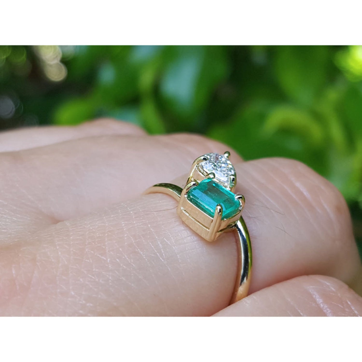 A yellow gold ring with a rectangular emerald-cut emerald and a pear cut diamond, shown worn on a person's finger against a blurred green background.