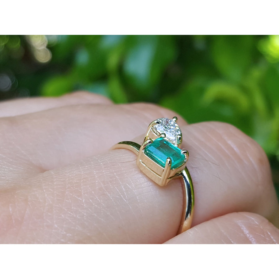 A yellow gold ring with a rectangular emerald-cut emerald and a pear cut diamond, shown worn on a person's finger against a blurred green background.