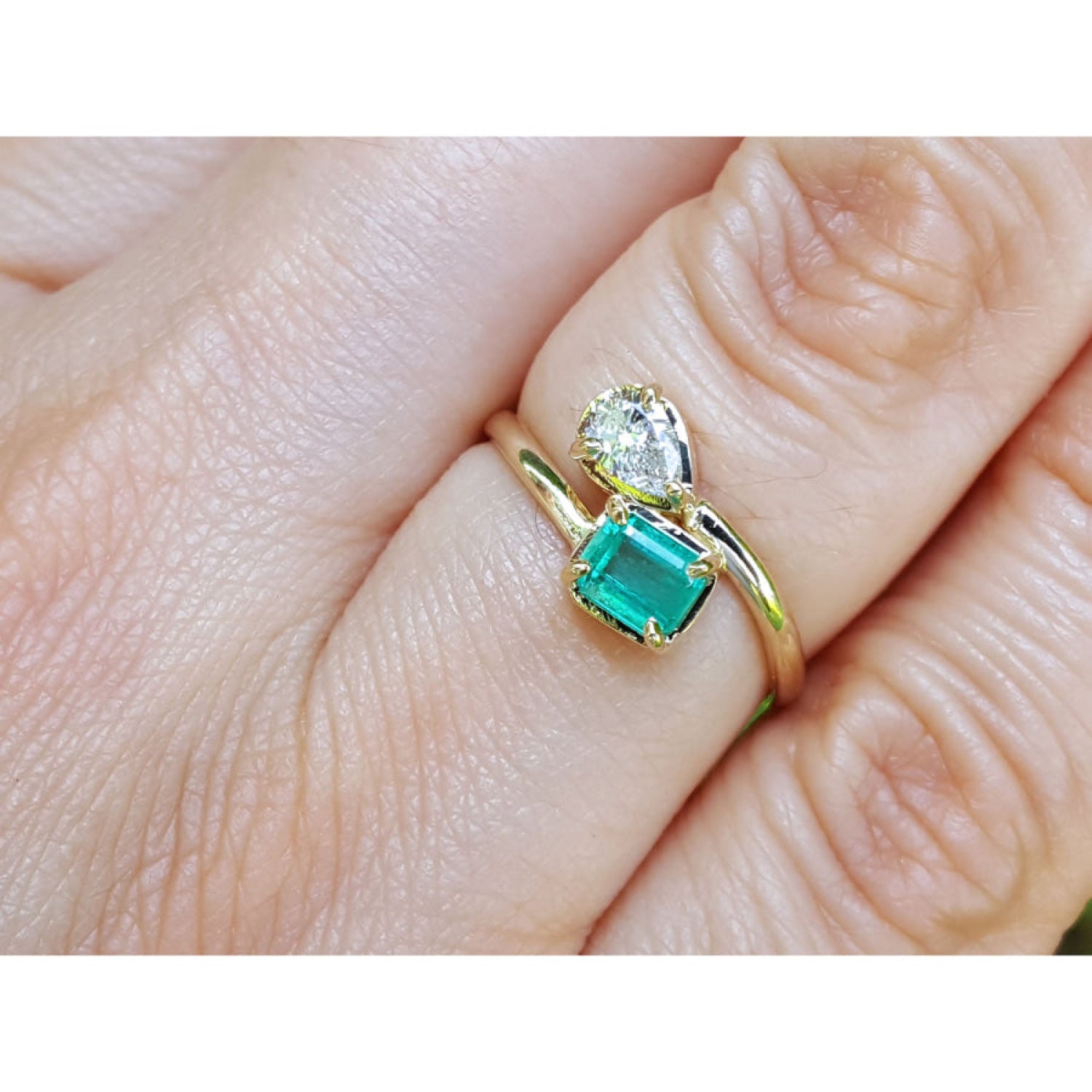 A close-up of a gold you and me ring with a square emerald-cut emerald and a smaller diamond, worn on a finger.