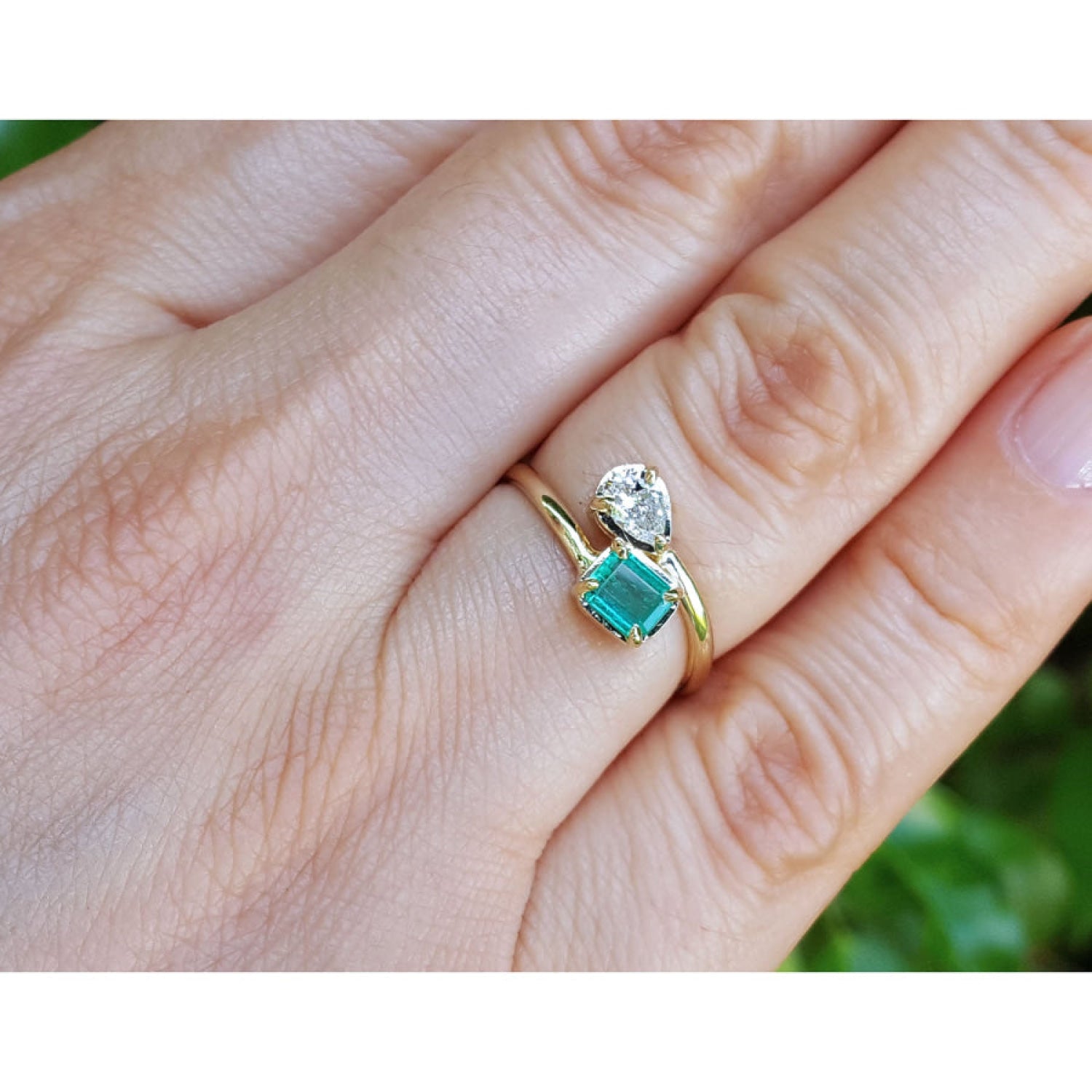 A gold ring with a square-cut emerald gemstone and a small pear cut diamond, shown worn on a person's finger.