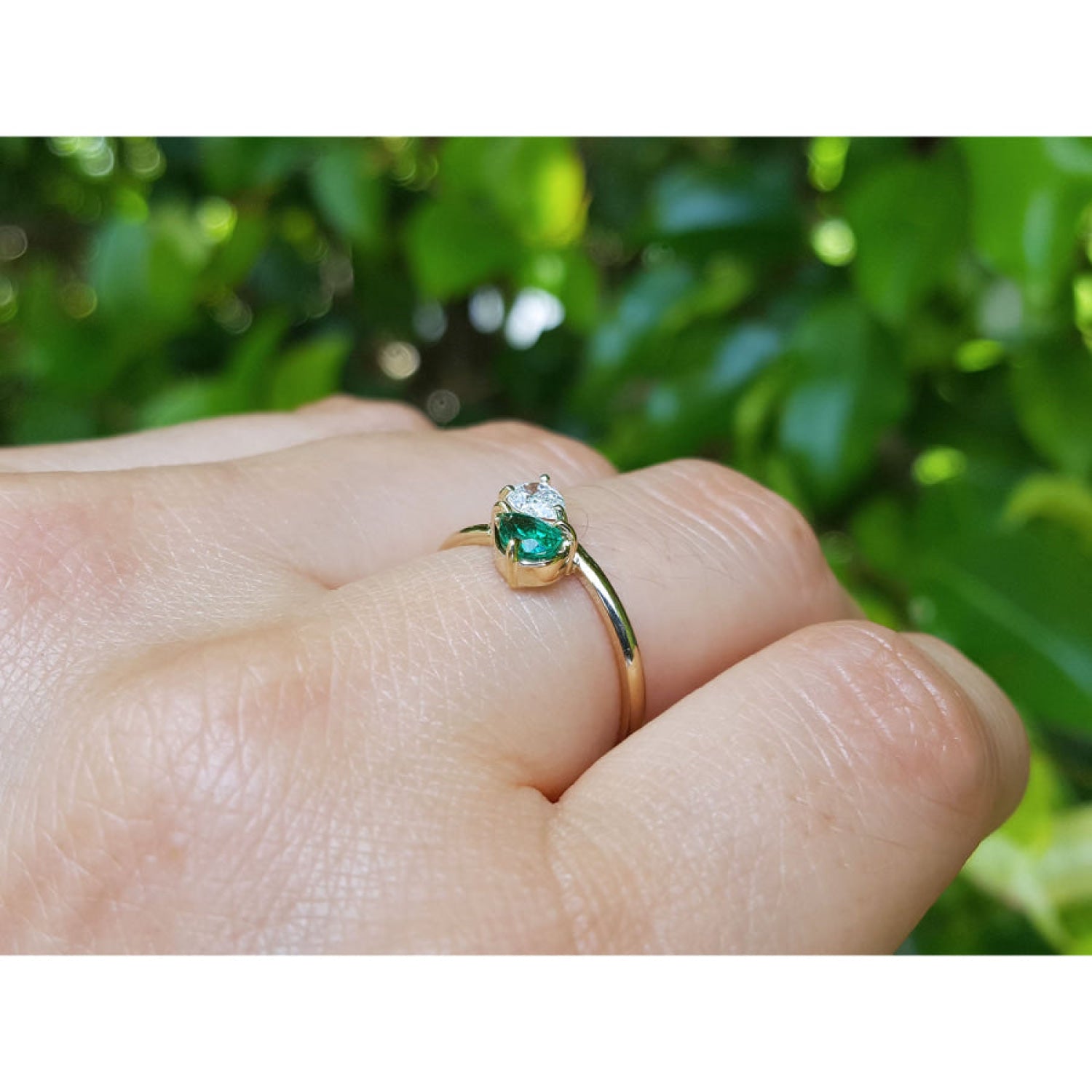 A gold ring with a pear-cut natural emerald, worn on a person's finger against a blurred background of green foliage.