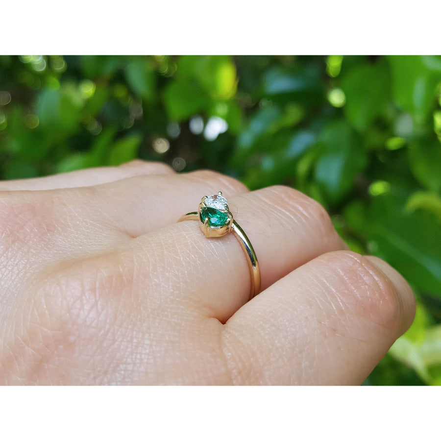 A gold ring with a pear-cut natural emerald, worn on a person's finger against a blurred background of green foliage.