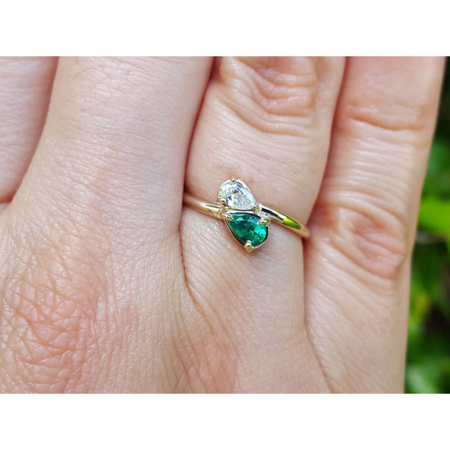 A gold ring with a diamond pear-cut gemstone and a natural emerald gemstone, shown worn on a person's finger.